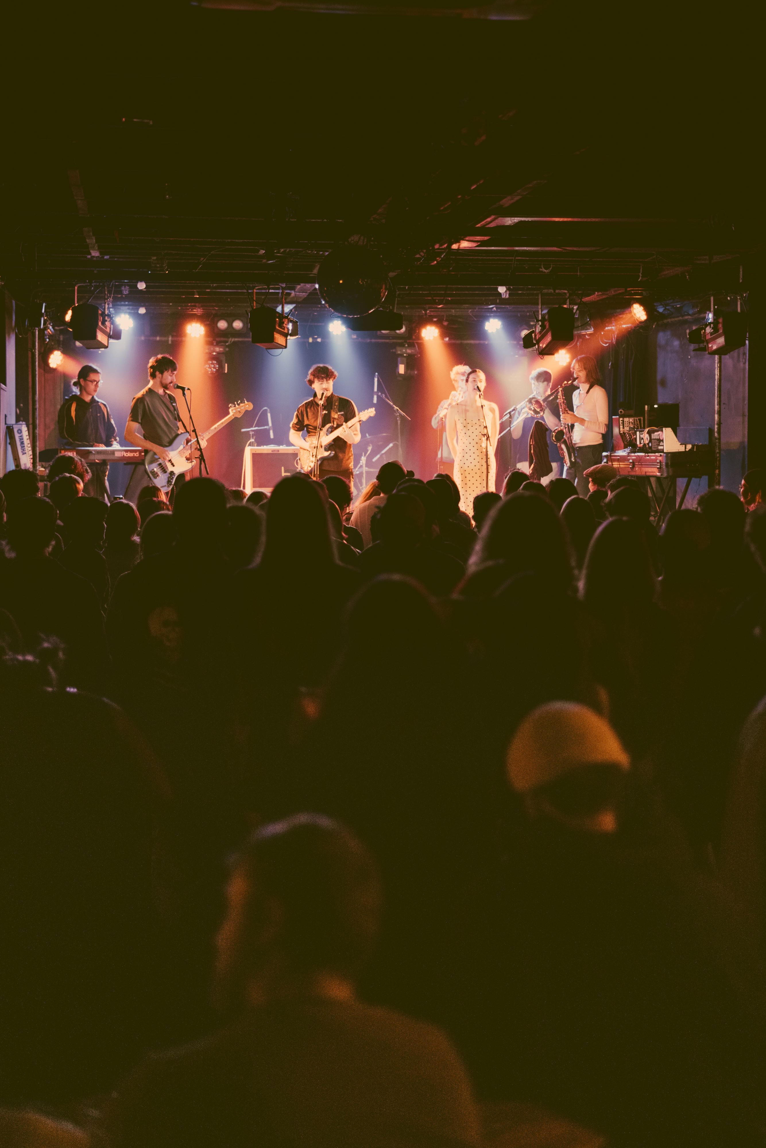 A shot taken from the back of a crowd, a large band is on stage lit under yellow and white lights